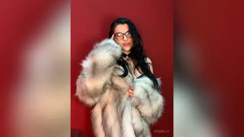 Glamorous woman in a luxurious white faux fur coat and glasses posing against a bold red background.