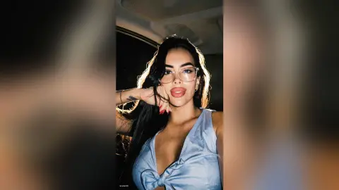 Glamorous woman with long dark hair and glasses wearing a blue satin top inside a car at night.