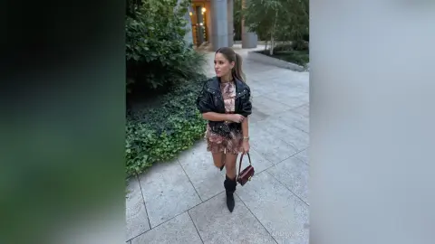 Stylish woman in a black leather jacket and floral mini dress walking on a stone path in a lush garden setting.