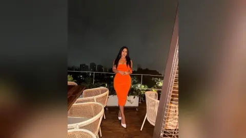 Elegant woman in a bright orange midi dress posing on a rooftop terrace at night with city lights background.