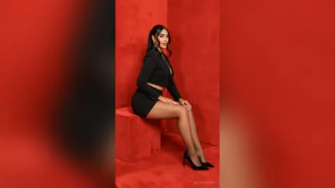 AllishaCoral — Elegant woman in a chic black outfit sitting against a vibrant red background in a sophisticated studio setting.