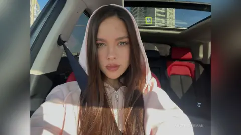 Young woman in a pink hoodie sitting in a car with sunlight creating a relaxed, casual mood.