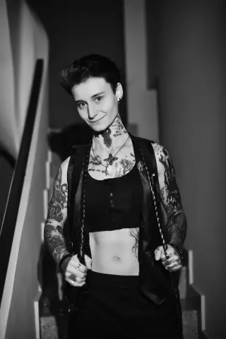 Smiling tattooed woman in a black crop top and vest posing in a moody indoor setting.