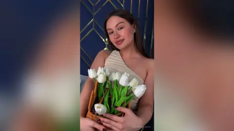 Smiling young woman in a cream halter top holding a bouquet of white tulips in a modern blue room.