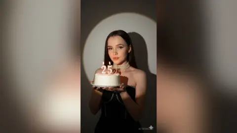 Young woman in a black dress holding a birthday cake with a 25 candle in a moody, dimly lit setting.