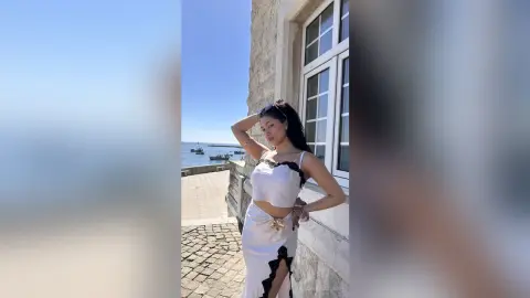 Stylish woman in a white satin crop top and lace skirt posing by a stone building overlooking the ocean.
