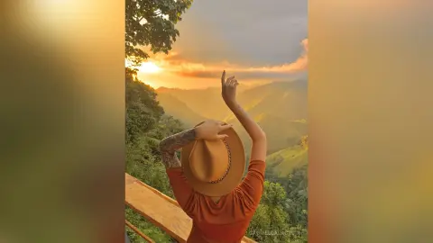 Woman in a brown shirt and sun hat enjoying a golden sunset view over lush green mountains.