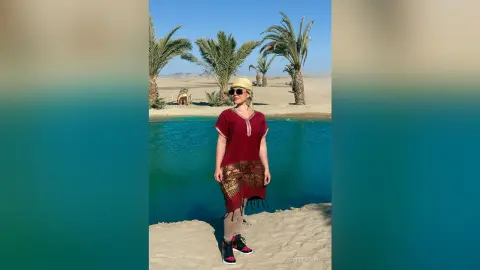 Woman in red tunic and patterned skirt standing by a desert oasis with palm trees under a sunny sky.