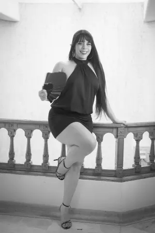 Smiling woman in a black halter dress holding a fan while leaning on a balcony railing in a black and white photo.