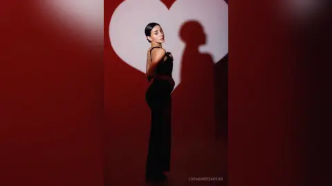 Elegant woman in a black evening gown posing against a red heart backdrop for a romantic mood.