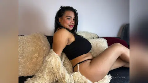 Smiling woman in black crop top and bikini relaxing on a cozy white faux fur rug in a bright bedroom.