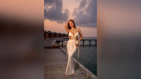 Elie poses on a wooden pier at sunset, wearing a white string bikini top and matching long skirt, with her hands in her hair.