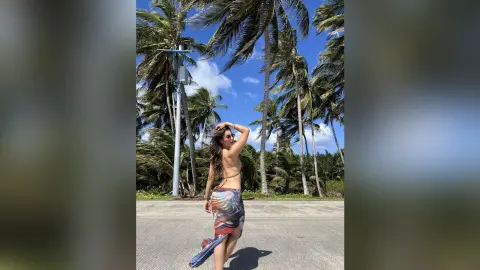 Woman in a colorful patterned sarong standing on a sunny tropical beach with palm trees under a blue sky.