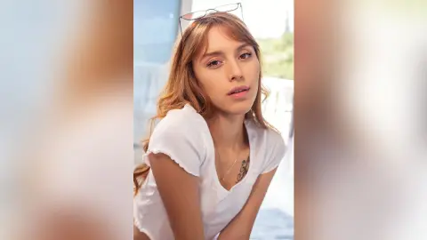 ValentinaGarcez wears a white t-shirt and glasses while looking at the camera with her long blonde hair down.
