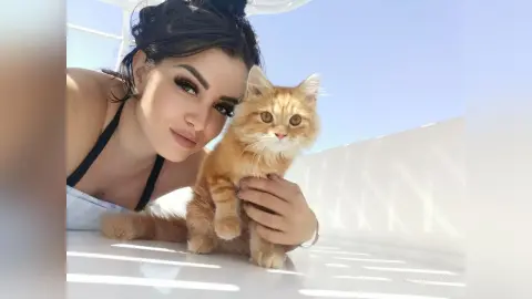 AnnaBanks poses outdoors in the sunlight, holding a fluffy ginger cat close to her face while wearing a black strap top.