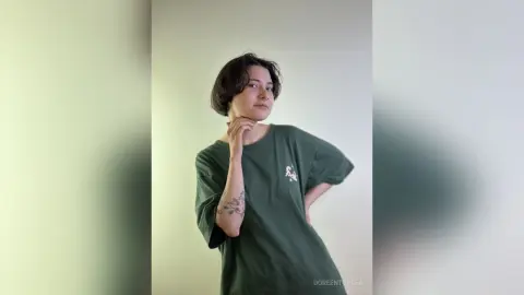 Young woman with short dark hair wearing a green oversized t-shirt posing in a minimalist indoor setting.