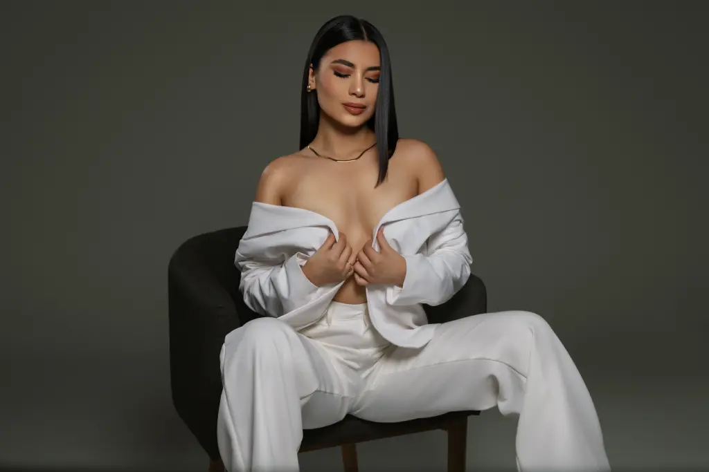 GabrielaLuviani poses in a black lace bra and black stockings, leaning back in a white chair with her long dark hair down.