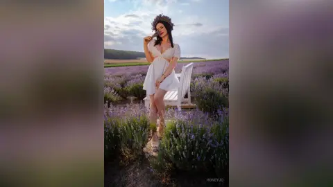 Dreamy woman in a white sundress wearing a flower crown sitting in a blooming lavender field at sunset.