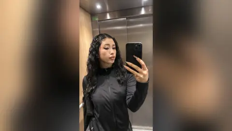 Young woman with long wavy hair wearing a black athletic zip-up jacket taking a selfie in an elevator.