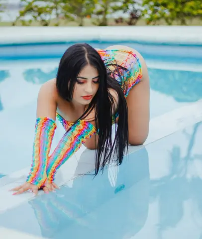 Brunette woman in colorful rainbow swimwear posing by a bright blue swimming pool on a sunny day.