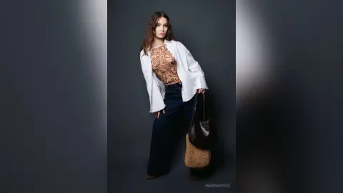 Young woman in a patterned blouse, white shirt, and dark flared jeans posing in a studio setting.