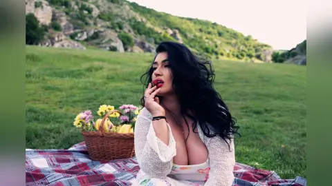 Woman in white lace outfit enjoying a romantic picnic with fruit basket in a lush green meadow.