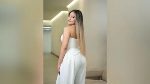 Elegant woman in a white strapless jumpsuit posing in a modern minimalist hallway with soft lighting.