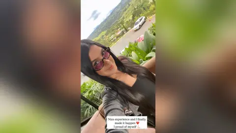 Smiling woman in sunglasses and black top enjoying a sunny outdoor view near lush green mountains.