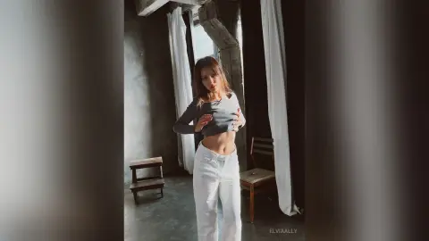 Young woman in a grey long-sleeve crop top and white jeans posing in a moody, minimalist concrete room.