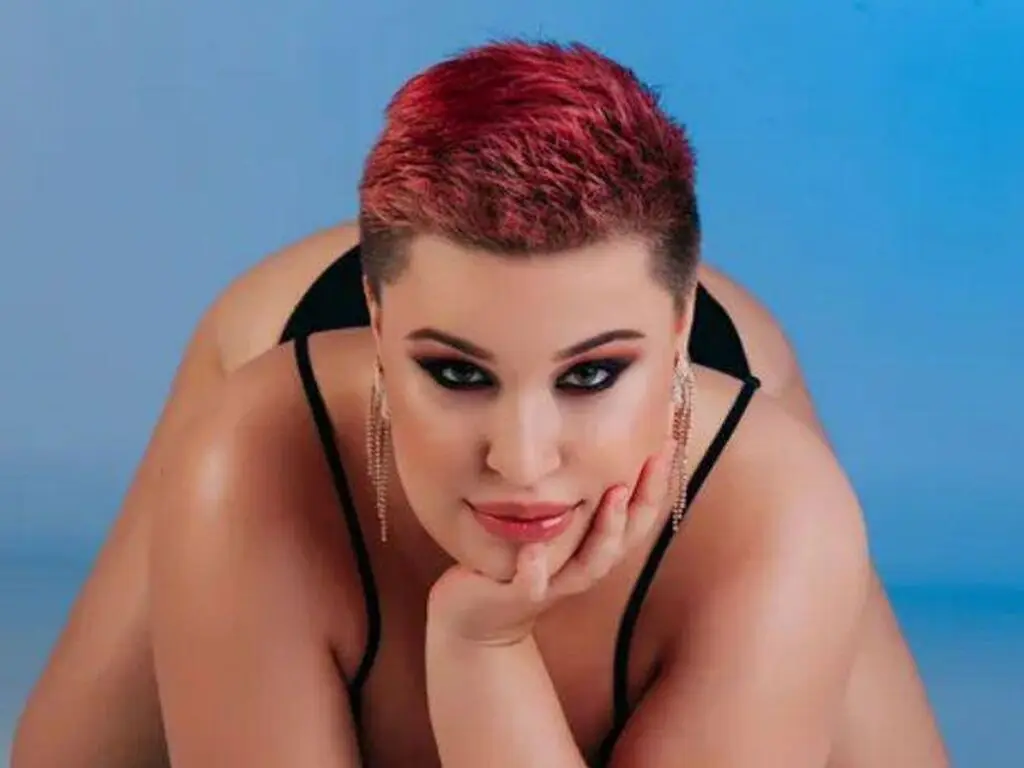 Curvy woman with short red hair and glamorous makeup posing in a black outfit against a blue background.