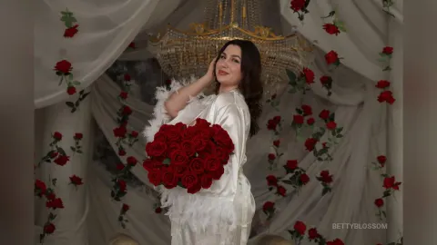 BettyBlossom — BettyBlossom poses in a white silk robe while holding a large bouquet of red roses against a draped backdrop.