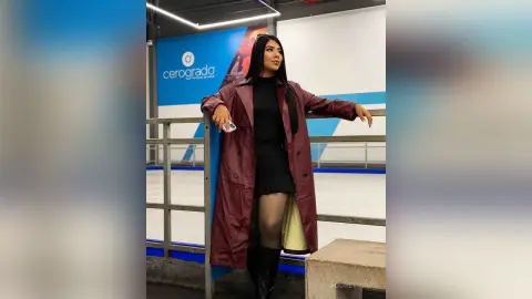 Stylish woman in a long burgundy coat and black dress standing in a modern indoor ice skating rink.