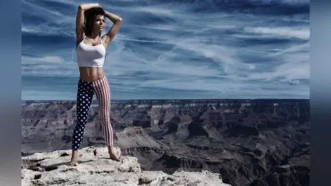 Woman in patriotic American flag leggings and white crop top standing on a Grand Canyon cliff under a cloudy sky.