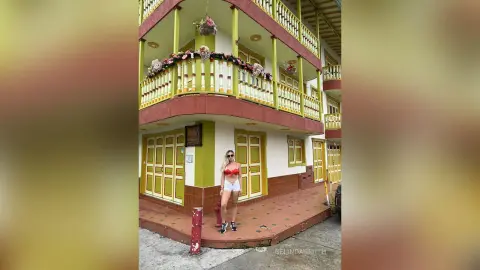 Young woman in a red bikini top and white shorts standing on a sunny balcony of a colorful tropical building.