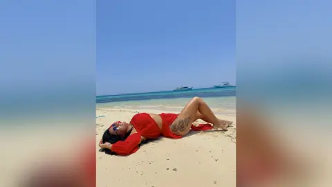 MissyJolie lies on her back on a sandy beach wearing a red bikini top and matching bottoms.
