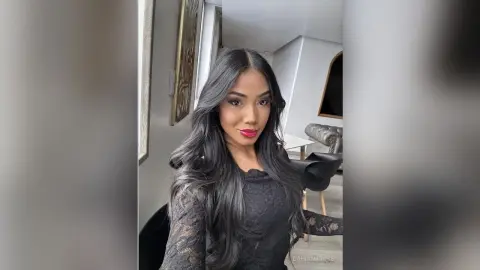 Young woman with long wavy dark hair wearing a black lace top in a bright modern living room.