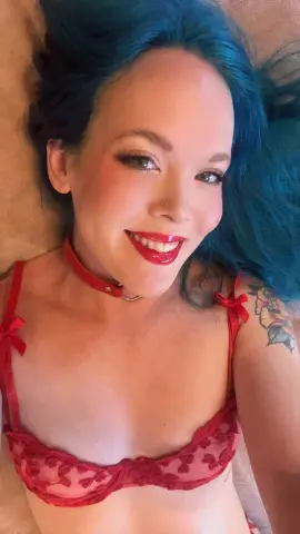 TemptingTiny smiles with bright red lipstick, wearing a red lace bra and a red choker, showing off her blue hair and shoulder tattoo.