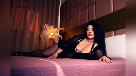 AdiraMaeven poses on a bed wearing a sheer black long-sleeve bodysuit that shows her black hair and cleavage.