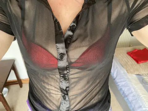 Woman in a sheer black button-down shirt and red bra posing in a bright bedroom setting.