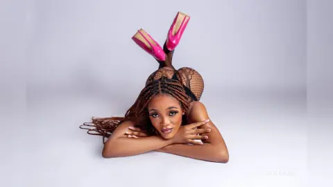 Beautiful woman with long braids in black fishnet lingerie and pink high heels posing on a white studio floor.