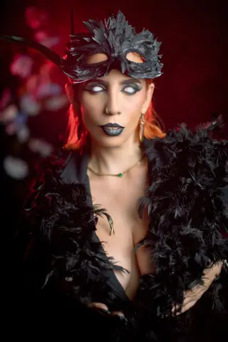 AdelinaVanz wears a black feathered mask and outfit, posing with dark lipstick and white eyes against a red background.
