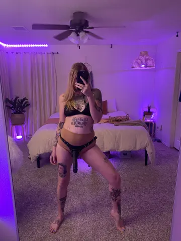 Tattooed woman in black lingerie posing in a purple lit bedroom with a cozy aesthetic.