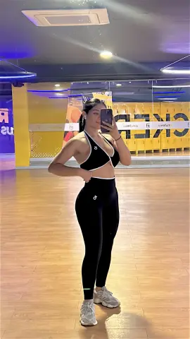 Athletic woman in black sports bra and leggings posing in a bright fitness studio gym setting.