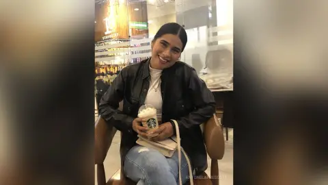 Smiling young woman in a black leather jacket holding a Starbucks coffee in a modern cafe setting.