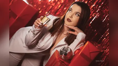 Glamorous woman in a white suit holding a champagne glass amidst red gift boxes and disco ball decor.
