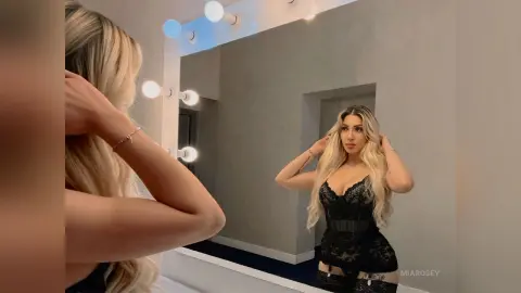 Blonde woman in a black lace corset outfit posing in a bright modern dressing room mirror.