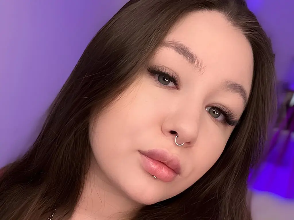 VanessaWeiss looks directly at the camera with long brown hair and a nose ring, wearing heavy eyelashes and lip gloss against a purple background.