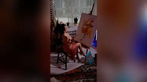 Woman in a pink dress painting an abstract landscape on a canvas in a bright, cozy studio setting.