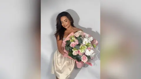 Smiling woman in a champagne satin dress holding a large bouquet of pink and white roses against a white wall.