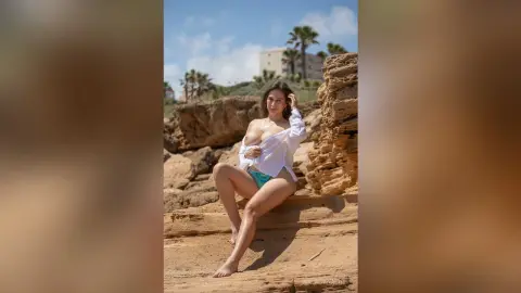 NataliaNacher poses outdoors on a rocky beach wearing a white open shirt and a light blue bikini bottom, showing off her cleavage and bare skin.